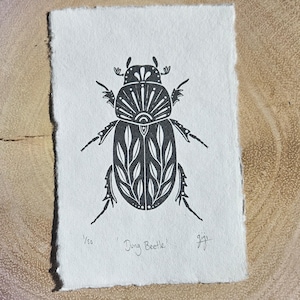 May include: A black and white linocut print of a dung beetle, featuring detailed patterns on its body and wings. The print is on a textured, rectangular paper with rough edges. The text "Dung Beetle" and "Gigi" are visible.