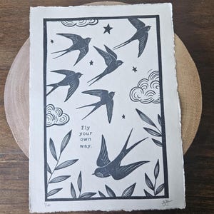 May include: A monochrome linocut print depicting swallows, stars, clouds, and leaves. The print includes the text "Fly your own way." The artwork is on textured paper with a deckled edge.