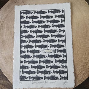 May include: A black and white linocut print on textured white paper, featuring a school of fish swimming in one direction, with a single fish swimming the opposite way. The print includes the text "Swim against the Current".