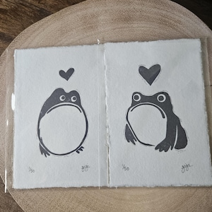 May include: Two black and white prints of a frog with a heart above it. The prints are on white paper and have the number 1/50 and the name gigi written on them.