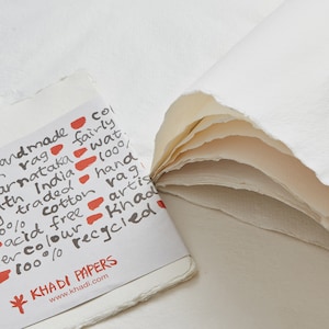 Pode incluir: Close-up de pap&eacute;is Khadi feitos &agrave; m&atilde;o. A imagem mostra uma pilha de papel branco com bordas &aacute;speras e um pequeno livreto com texto em preto e vermelho. O texto inclui detalhes sobre a composi&ccedil;&atilde;o e a origem do papel.
