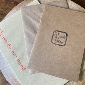 Pode incluir: Um conjunto de cart&otilde;es de felicita&ccedil;&otilde;es e envelopes de papel kraft. Um cart&atilde;o tem um carimbo preto "Thank You" numa moldura decorativa. Outro cart&atilde;o diz "please do not bend" em vermelho. Os cart&otilde;es est&atilde;o sobre um prato decorativo.