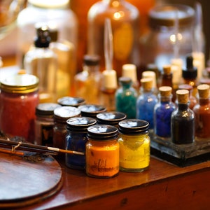 Pode incluir: Uma cole&ccedil;&atilde;o de materiais de arte, incluindo potes de tinta em v&aacute;rias cores, pequenas garrafas de vidro com rolhas e pinc&eacute;is. Os potes de tinta s&atilde;o em tons de laranja, amarelo e azul. Uma paleta de madeira tamb&eacute;m &eacute; vis&iacute;vel.