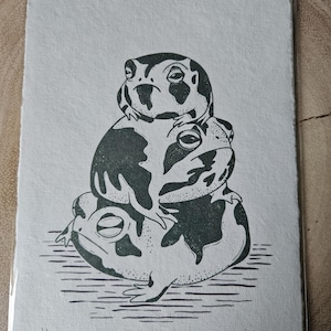 Może przedstawiać: Czarno-biała linorytnica przedstawiająca trzy żaby ułożone jedna na drugiej. Żaby są narysowane w prosty, stylizowany sposób. Druk jest podpisany "gigi" i ponumerowany "1/50".