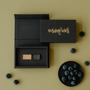 Op de afbeelding: Een zwarte doos met het woord "herinneringen" in gouden letters. De doos is open en bevat een kleine gouden en transparante USB-stick. De doos staat op een groene ondergrond met verspreide blauwe bessen.