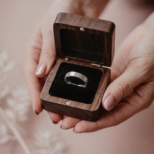 Può includere: Un anello d'argento esposto in un portagioie di legno scuro aperto, con interno in velluto nero. L'anello è al centro, tenuto da mani con unghie curate. L'anello ha una fascia piatta.