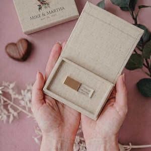 Puede incluir: Una caja de textura de lino beige que contiene una unidad flash USB metálica grabada con el texto Happy 7th Anniversary 2023. La tapa a juego presenta una ilustración de un pájaro y el texto MIKE & MARTHA MAY 26, 2023. La caja tiene bisagras.