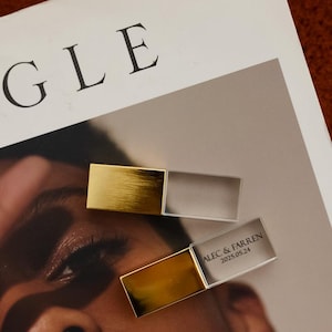 Può includere: Due chiavette USB rettangolari, dorate e argentate, con finitura in metallo spazzolato. Una chiavetta è aperta, rivelando il testo "ALEC & FARREN 2025.05.24". Le chiavette sono su una rivista con la parola "VOGUE".