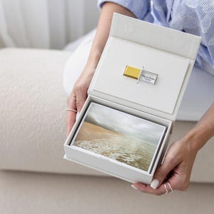 Può includere: Una scatola di presentazione rettangolare bianca contenente una pila di stampe fotografiche rettangolari di una spiaggia e una chiavetta USB dorata e argentata fissata al coperchio interno, con il testo inciso "Kay and Andrew 12.05.2023".