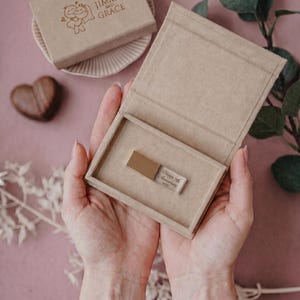 May include: A rectangular tan cardboard box containing a metallic USB flash drive engraved with the text Happy 7th Anniversary 2022. A matching tan box lid with the printed text Jimmy and Grace sits nearby on a small ridged plate.
