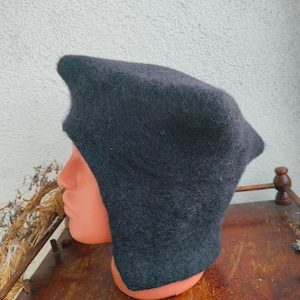 May include: A dark gray, hooded hat with ear flaps. The hat is made of a soft, felted material and is displayed on a mannequin head. The hat has a pointed top and covers the ears and neck.