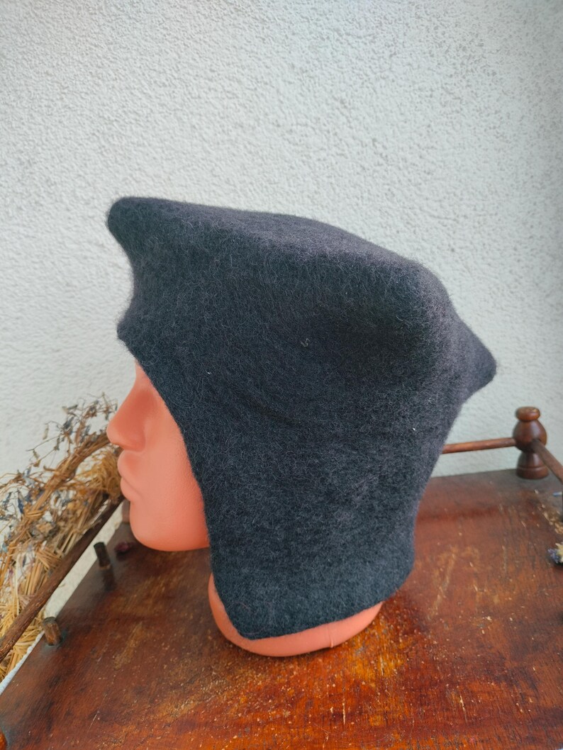 May include: A dark gray, hooded hat with ear flaps. The hat is made of a soft, felted material and is displayed on a mannequin head. The hat has a pointed top and covers the ears and neck.