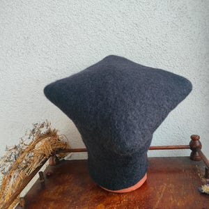 May include: A dark gray felted hat with a unique, angular shape. The hat is displayed on a wooden stand with a reddish-brown base. The background is a light-colored wall.
