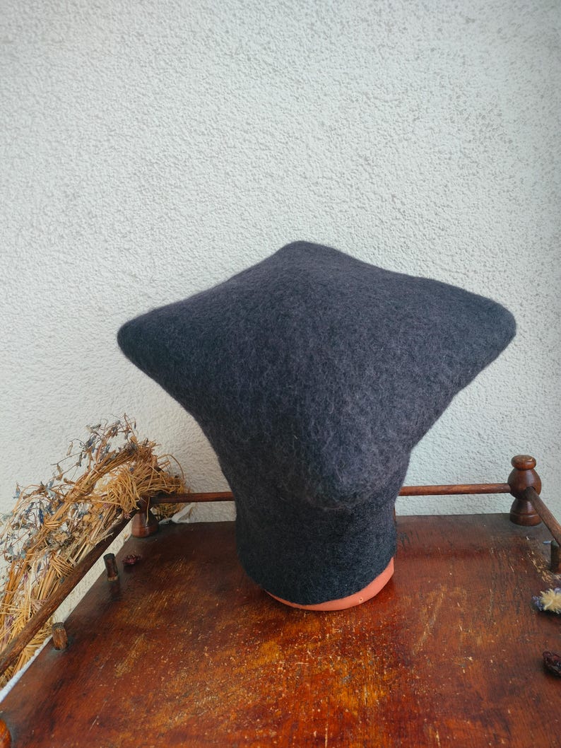 May include: A dark gray felted hat with a unique, angular shape. The hat is displayed on a wooden stand with a reddish-brown base. The background is a light-colored wall.