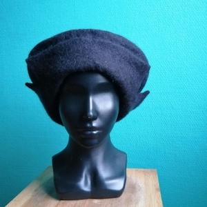 Peut inclure: Un béret en laine feutrée gris anthracite foncé avec une calotte arrondie structurée et un large bord replié qui s'étend légèrement sur les côtés. Le chapeau est présenté sur une tête de mannequin noire lisse, mettant en valeur la matière dense.