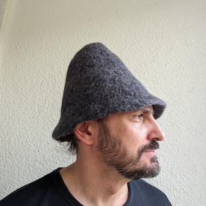 Peut inclure: Un chapeau en laine feutrée gris foncé doté d'une calotte conique haute et d'un bord court légèrement évasé. La matière présente un aspect texturé et chiné avec des fibres visibles sur toute la surface, offrant un rendu dense et uniforme.