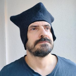 May include: A dark navy blue hat with a unique, angular design. The hat has a pointed top and covers the ears and forehead. The hat is made of a soft, textured material. The person in the image is wearing a dark blue v-neck shirt.