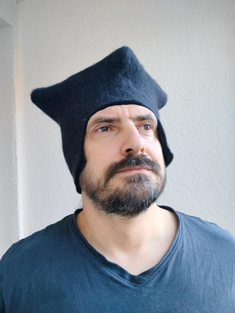 May include: A dark navy blue hat with a unique, angular design. The hat has a pointed top and covers the ears and forehead. The hat is made of a soft, textured material. The person in the image is wearing a dark blue v-neck shirt.
