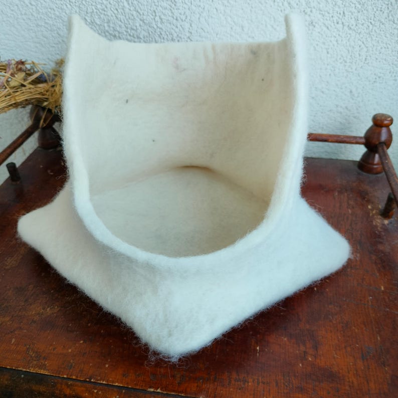 May include: A cream-colored, felted cat bed with a square base and curved, upright sides. The bed has a soft, textured appearance and is designed to provide a cozy space for a cat to rest. The bed is made of a thick, natural material.