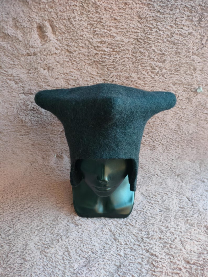 May include: A dark teal felt hat with a unique, angular design. The hat has a wide, flat brim and a tall, pointed crown. The hat is displayed on a dark green mannequin head.
