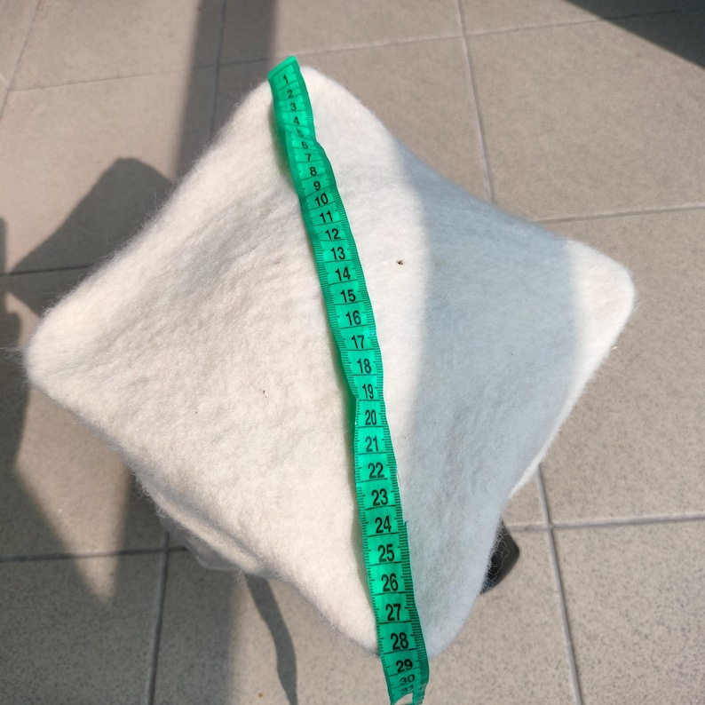 May include: A white, square-shaped felted hat is shown with a green measuring tape. The hat appears to be approximately 12 inches in length. The hat is made of a soft, textured material.