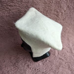 May include: A white, felted hat with a square crown and a slightly rounded brim. The hat is displayed on a black mannequin head against a textured, mauve background. The hat appears to be handmade.