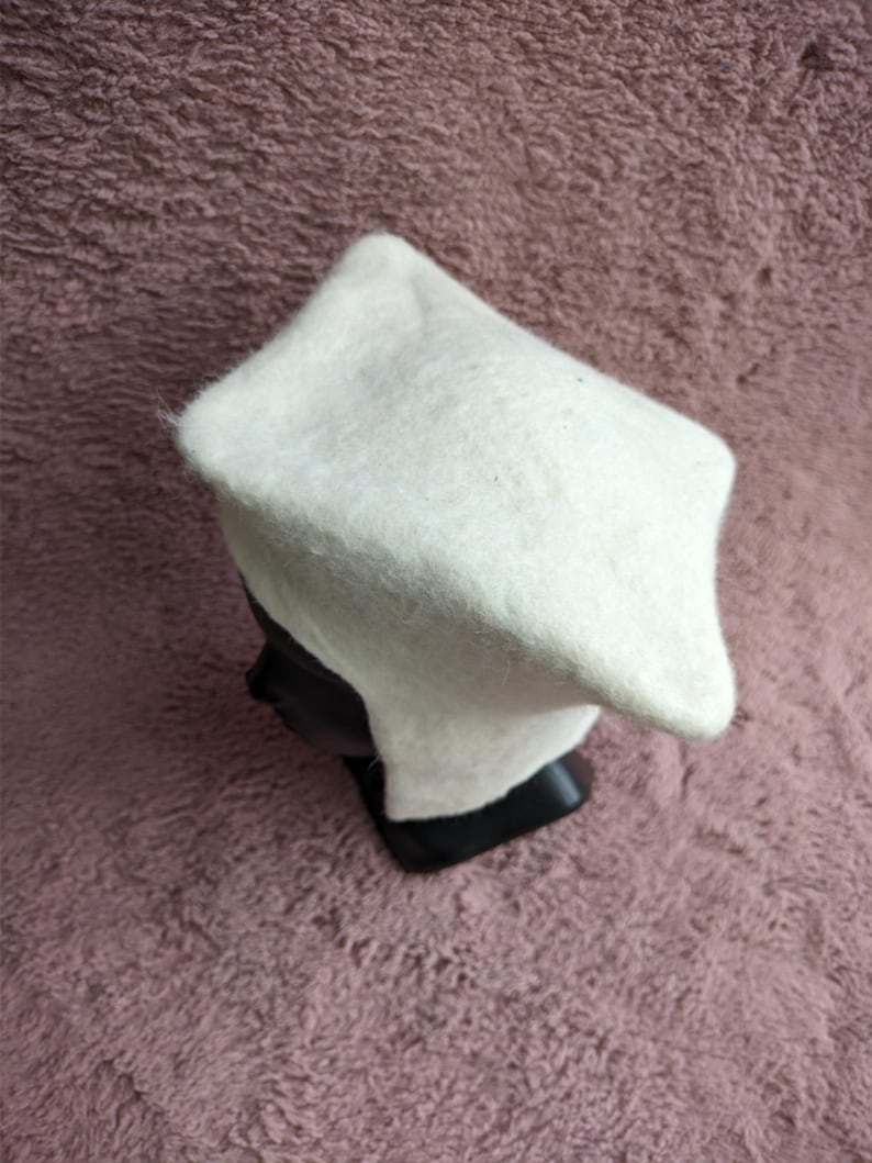 May include: A white, felted hat with a square crown and a slightly rounded brim. The hat is displayed on a black mannequin head against a textured, mauve background. The hat appears to be handmade.