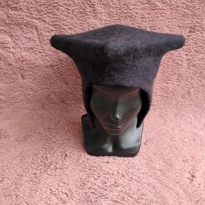 May include: A dark gray, felted hat with a distinctive, pointed shape. The hat has a brim that extends outward on each side. The hat is displayed on a black mannequin head against a textured, pink background.