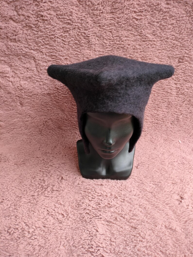 May include: A dark gray, felted hat with a distinctive, pointed shape. The hat has a brim that extends outward on each side. The hat is displayed on a black mannequin head against a textured, pink background.