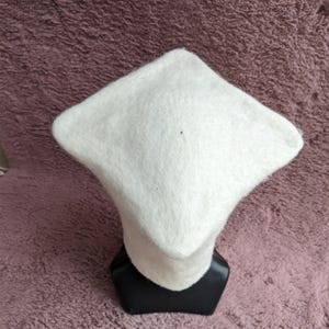 May include: A white, square-shaped hat with a rounded brim, displayed on a black mannequin head. The hat is made of a soft, textured material. The background is a textured, mauve-colored surface.