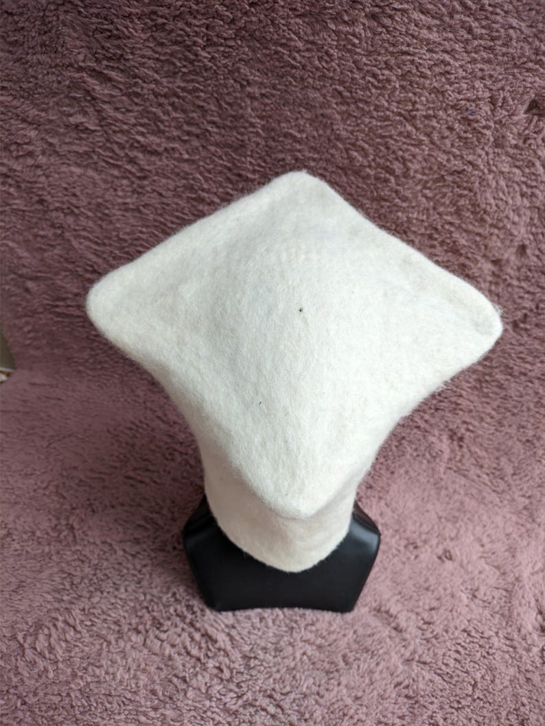 May include: A white, square-shaped hat with a rounded brim, displayed on a black mannequin head. The hat is made of a soft, textured material. The background is a textured, mauve-colored surface.
