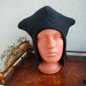May include: A dark gray, medieval-style hat with a distinctive pointed crown and ear flaps. The hat is made of a soft, felted material and is displayed on a mannequin head. The hat's design suggests historical or fantasy themes.