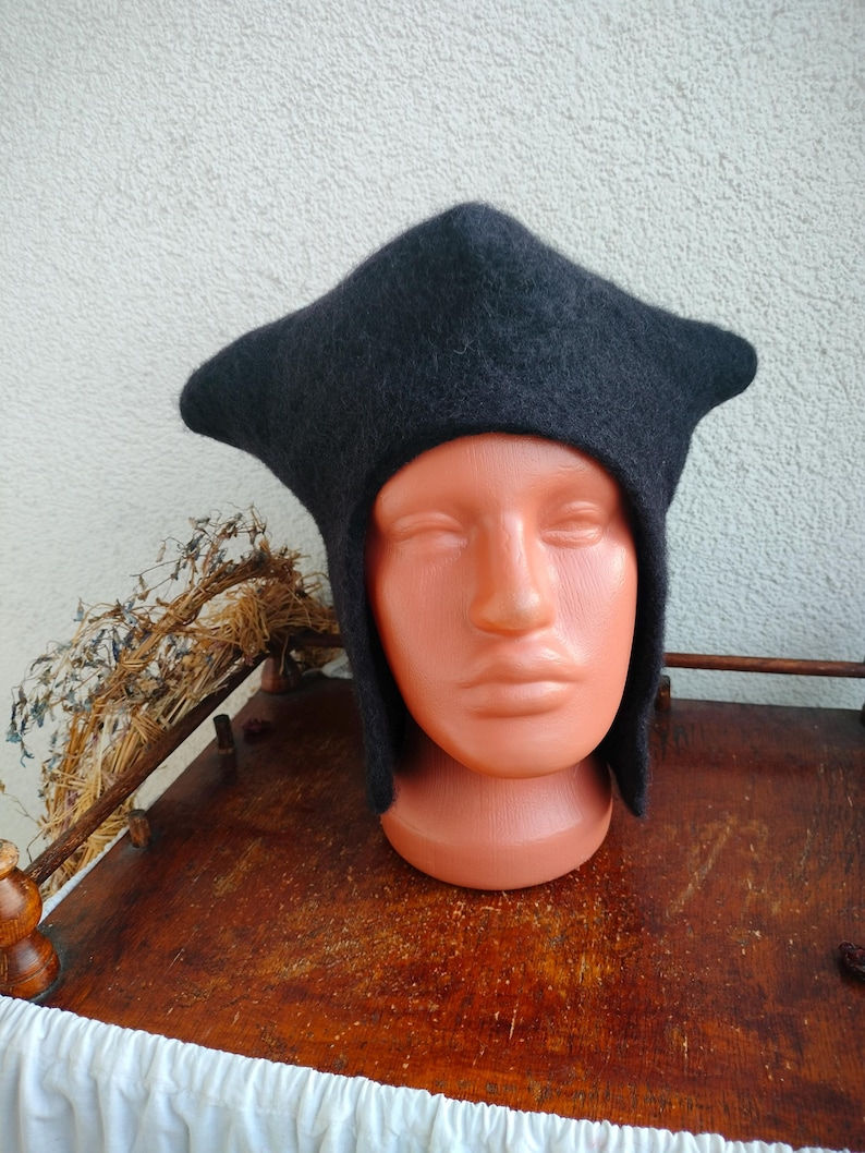 May include: A dark gray, medieval-style hat with a distinctive pointed crown and ear flaps. The hat is made of a soft, felted material and is displayed on a mannequin head. The hat's design suggests historical or fantasy themes.