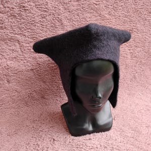 May include: A dark gray, hooded hat with a pointed crown and ear flaps. The hat is made of a felted material and is displayed on a black mannequin head against a pink textured background. The hat's design is reminiscent of medieval headwear.