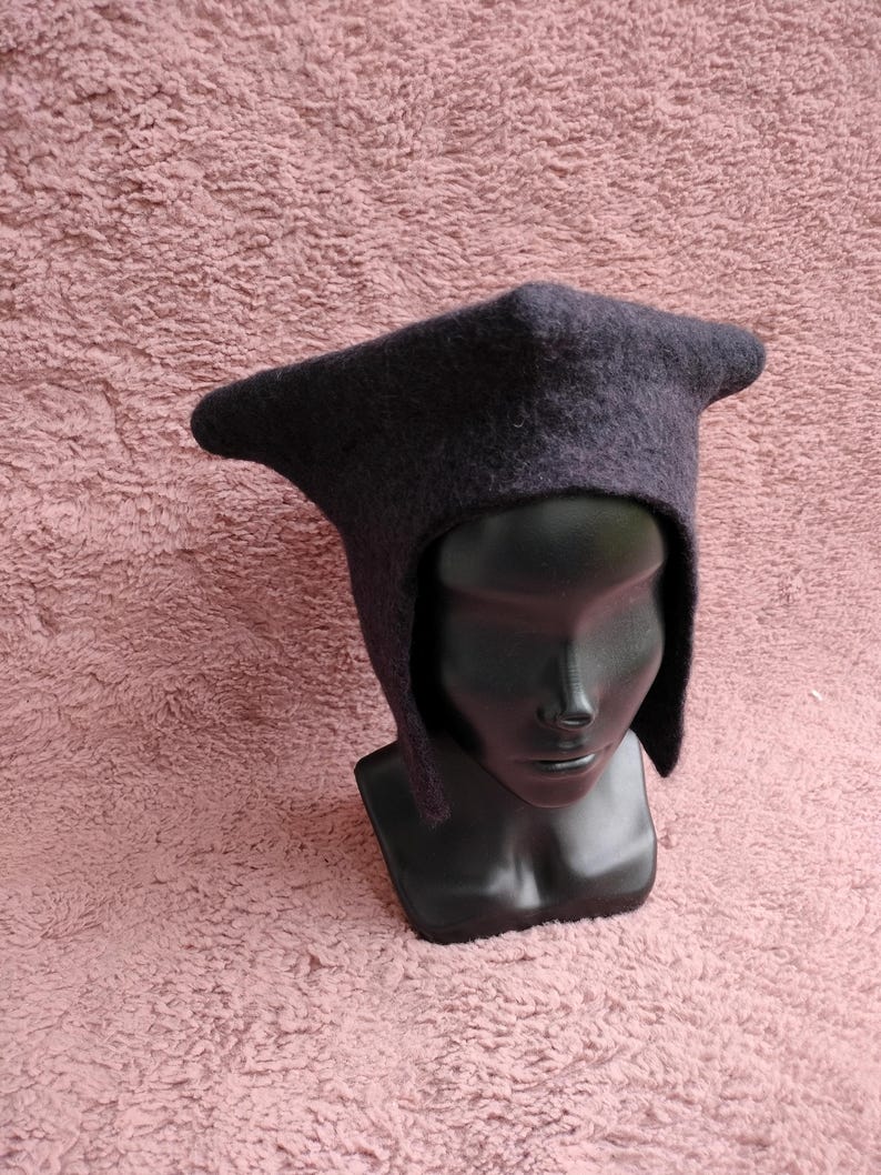 May include: A dark gray, hooded hat with a pointed crown and ear flaps. The hat is made of a felted material and is displayed on a black mannequin head against a pink textured background. The hat's design is reminiscent of medieval headwear.