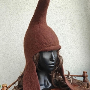 Può includere: Un cappello di feltro marrone con una corona alta e appuntita e paraorecchie. Il cappello è esposto su una testa di manichino nera con lunghi capelli castani ondulati. Il colore del cappello è un marrone intenso e terroso, e sembra essere fatto di un materiale morbido e strutturato.