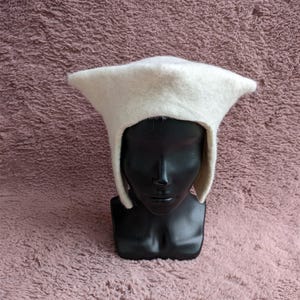 May include: A white felt hat with a unique, angular design. The hat features a pointed crown and ear flaps, displayed on a black mannequin head against a textured, mauve background. This hat is a distinctive accessory.