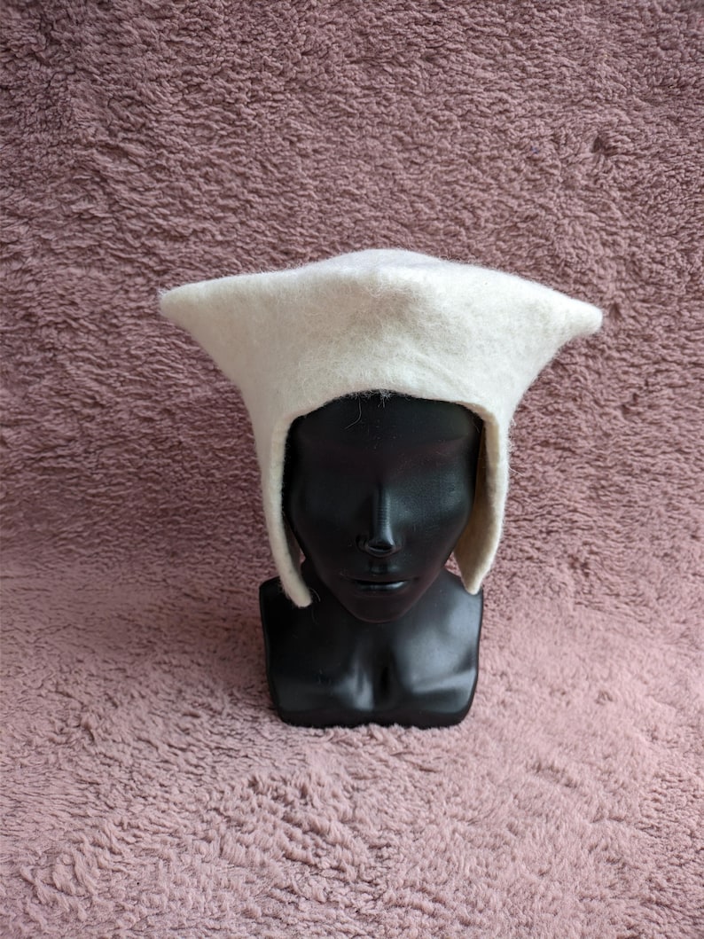 May include: A white felt hat with a unique, angular design. The hat features a pointed crown and ear flaps, displayed on a black mannequin head against a textured, mauve background. This hat is a distinctive accessory.