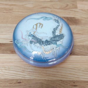 May include: A round, blue trinket box featuring a detailed dragon design. The dragon is rendered in shades of grey, gold, and blue, with intricate detailing. The box has a glossy finish and rests on a wooden surface.