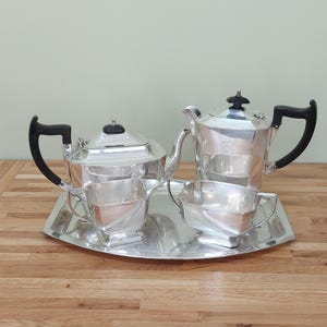 May include: A silver tea set comprising a tray, two teapots, and two cream jugs. The teapots feature black handles and lids. The set is presented on a wooden surface.