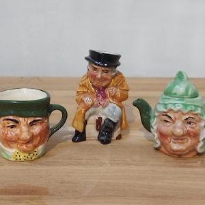 May include: Three ceramic figurines: a creamer, a seated man, and a teapot. The creamer has a green rim and handle, with a face design. The seated man wears a black top hat and tan coat. The teapot has a green lid and spout, with a face design.