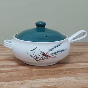 May include: A white ceramic soup tureen with a teal lid and handles. The tureen is decorated with a floral design in red, green, and blue. The tureen is sitting on a wooden surface.