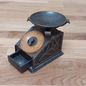 May include: An antique metal scale with a small, round bowl and a dial. The scale has a drawer and decorative floral accents. The dial is tan and the scale is dark grey. A vintage kitchen decor item.