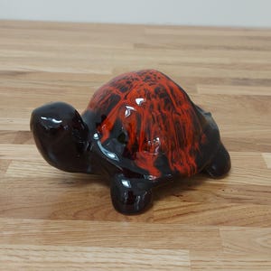 May include: A ceramic turtle ornament with a glossy finish. The turtle's shell features a vibrant red and black marbled pattern, while the head, legs, and body are solid black. The ornament is displayed on a wooden surface.