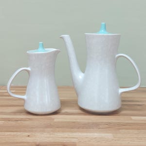 May include: A white ceramic coffee pot and creamer set with a speckled design. The coffee pot has a long spout and a handle, and both pieces have light blue lids. The set is displayed on a wooden surface.