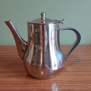 May include: A shiny stainless steel teapot with a curved spout and handle. The teapot has a lid with a small knob and a hinged handle. The teapot is on a wooden surface.