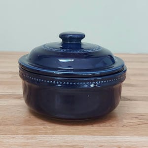 May include: A deep navy blue ceramic lidded bowl featuring a glossy finish and a decorative beaded texture along the rim of both the lid and the base. The lid includes a small, rounded knob handle centred on top of the circular container.