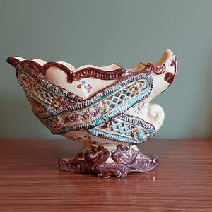 May include: A decorative ceramic vase with a cream base and detailed design. The vase has a lattice pattern with teal and brown accents, and floral motifs. The base and rim are dark brown. The vase is displayed on a wooden surface.
