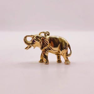 May include: A gold-coloured elephant pendant with a raised trunk and a small, dark-coloured gem in its eye. The pendant is detailed with textured skin and a loop for a chain. The elephant is standing on four legs.