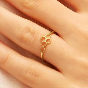 Könnte beinhalten: Ein goldfarbener Ring mit einem Wabenmuster. Der Ring ist zierlich und dünn, wobei sich das Wabenmuster über die Oberseite des Fingers erstreckt. Der Ring wird an einer Hand mit gepflegten Nägeln getragen.